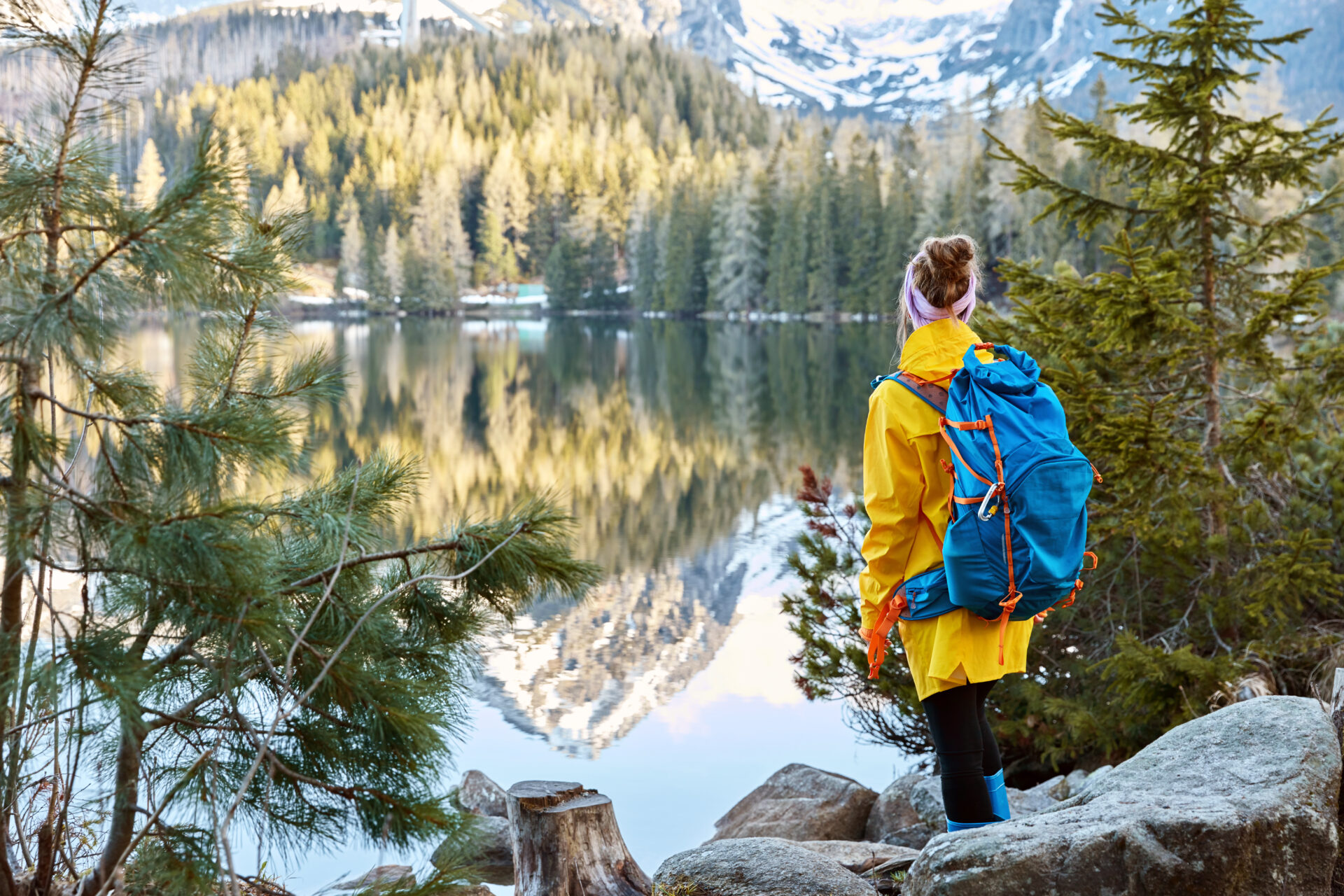 Horizontal view of female tourist enjoys tranquil remote mountain lake view, stands back to camera, carries big blue rucksack, wears raincoat and rubber boots, stands near green firs on stones